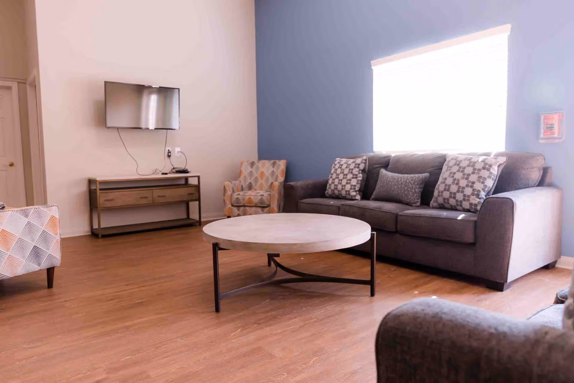 A bright living room with a gray sofa adorned with patterned pillows, two patterned armchairs, a round coffee table with a metal frame, a wall-mounted flat-screen TV above a wooden console table, and a large window with blinds letting in natural light.