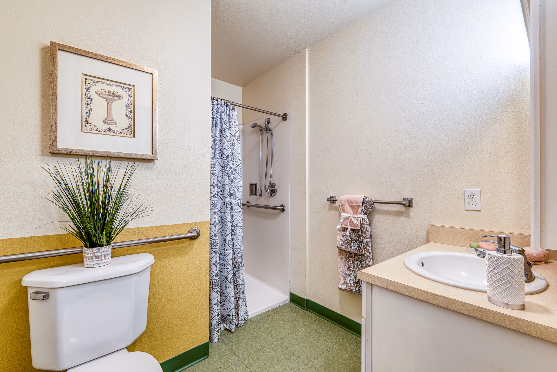 A bathroom with a white toilet topped with a potted plant, a framed picture on the wall, a shower with a patterned curtain, a towel rack holding folded towels, and a sink with a soap dispenser and soap bars on the countertop.