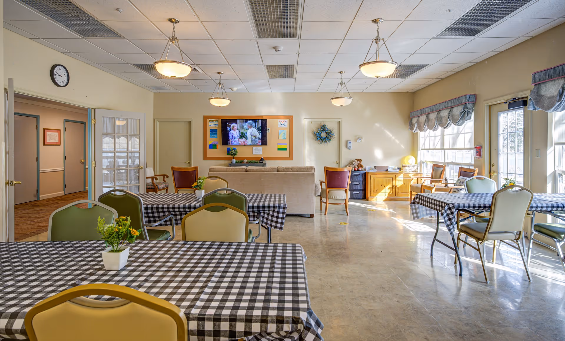 A bright and spacious common area with multiple tables covered in black and white checkered tablecloths, each with a small potted plant centerpiece. The room has several chairs in different colors around the tables. At the far end, there is a beige couch facing a wall-mounted TV displaying a show with two elderly people. The walls are light-colored, with a wreath and some decorations. Large windows on the right side let in natural light, and there are ceiling lights evenly spaced. Doors and a clock are visible on the left side of the room.