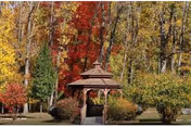 A wooden gazebo surrounded by trees with autumn foliage in vibrant red, yellow, and green colors, set in a grassy outdoor area.