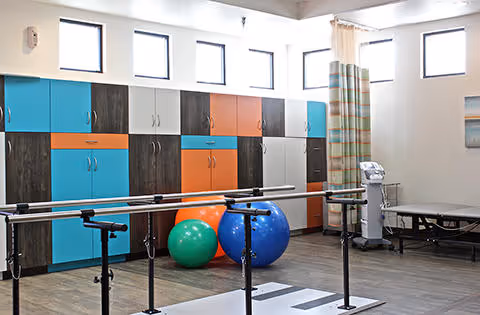 Physical therapy room with colorful storage cabinets, parallel bars for walking exercises, large exercise balls, a treatment table, and medical equipment, all in a well-lit space with multiple windows.