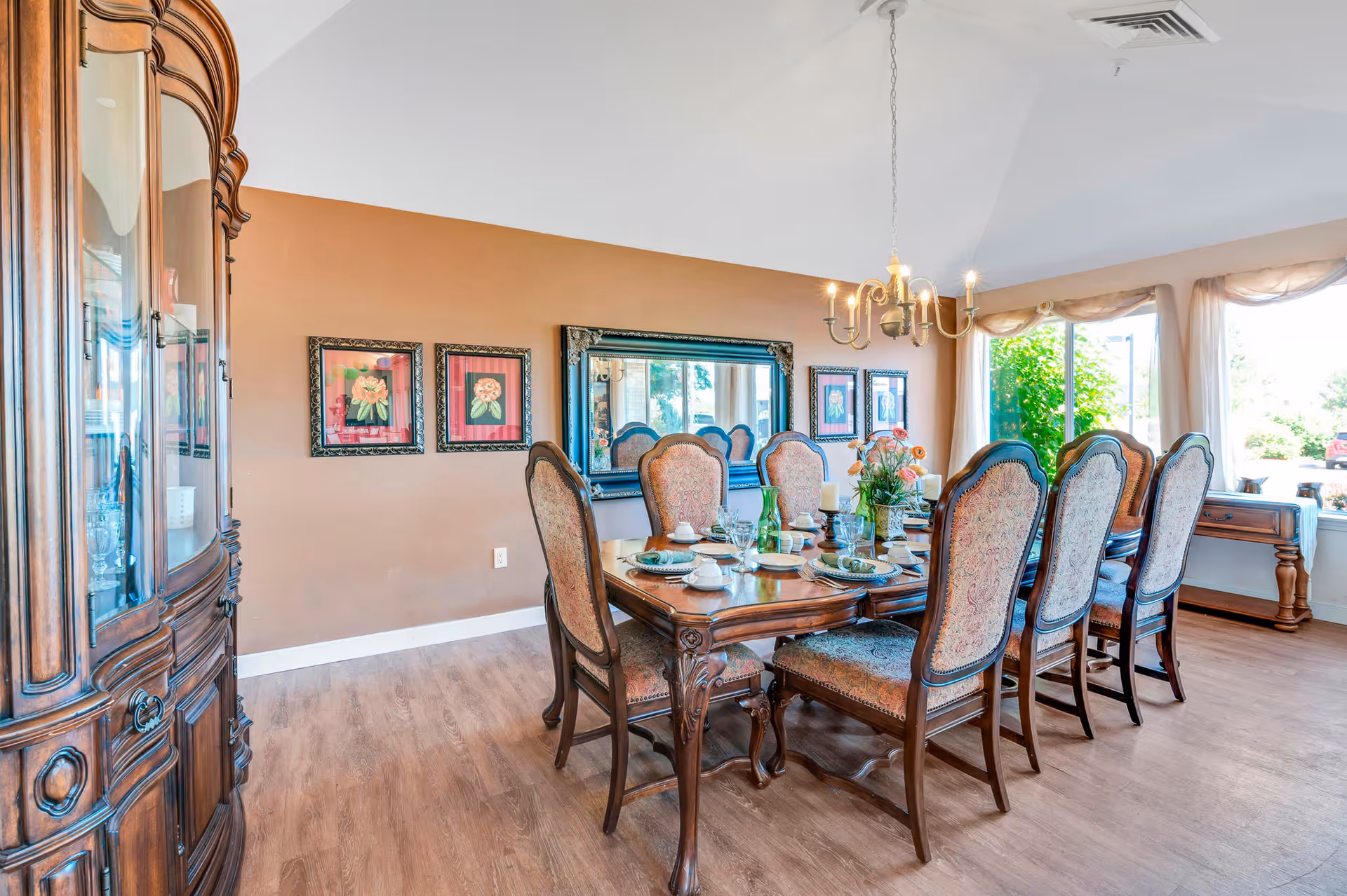 A well-lit dining room with a large wooden dining table set for eight people. The chairs have ornate wooden frames and upholstered backs and seats. On the table are plates, glasses, napkins, and a floral centerpiece. The room has a wooden floor, a chandelier hanging above the table, a large mirror on the wall, and three framed floral paintings. Large windows with sheer curtains allow natural light to fill the space.