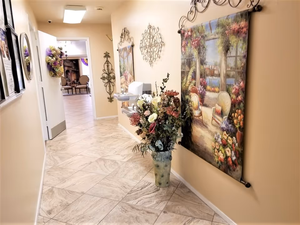 Decorated hallway inside a senior living facility with tiled floor, wall art, and a large floral arrangement.