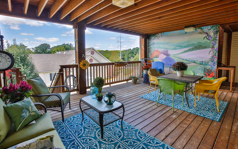 Covered outdoor patio area with wooden flooring and ceiling beams, furnished with a green cushioned sofa, a glass-top coffee table, colorful chairs around a dining table on a blue patterned rug, potted plants, and a mural of a scenic landscape on one wall.