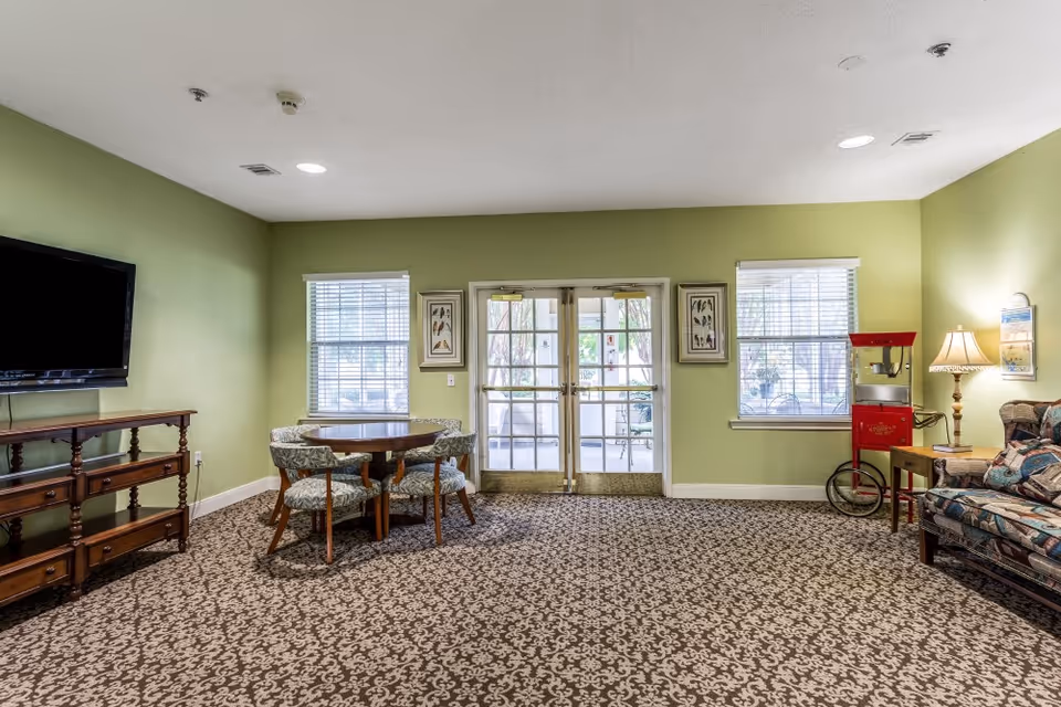 A common area in Hearthstone Assisted Living with green walls, patterned carpet, a round wooden table with four upholstered chairs, a flat-screen TV on a wooden stand, a patterned sofa, a side table with a lamp, a red popcorn machine, and double glass doors leading outside.
