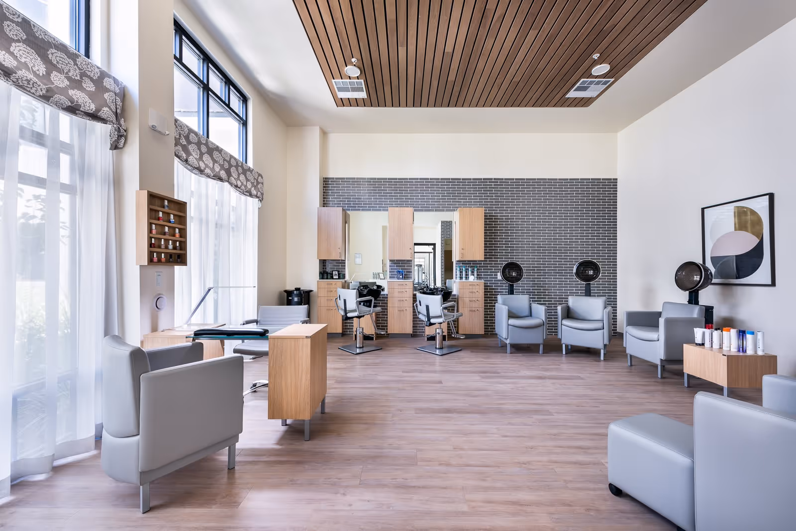 A bright and modern salon area with large windows covered by sheer white curtains and patterned valances. The room features light wood flooring, several gray salon chairs, hair drying stations, and a wall with mirrors and wooden cabinets. There is also a small table with various hair products and a framed abstract artwork on the wall.