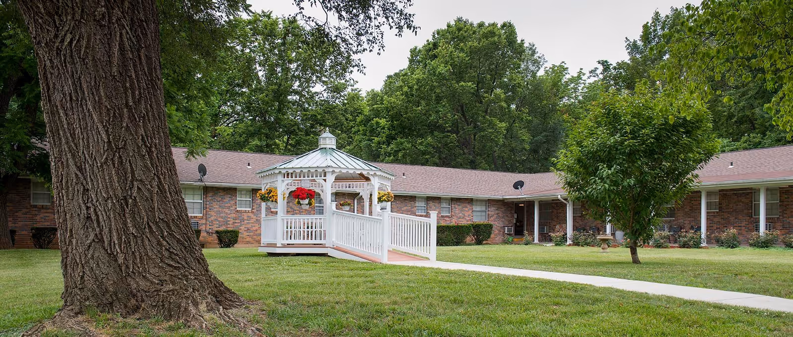 Single-story brick senior living building with a white gazebo on a grassy courtyard and large trees.