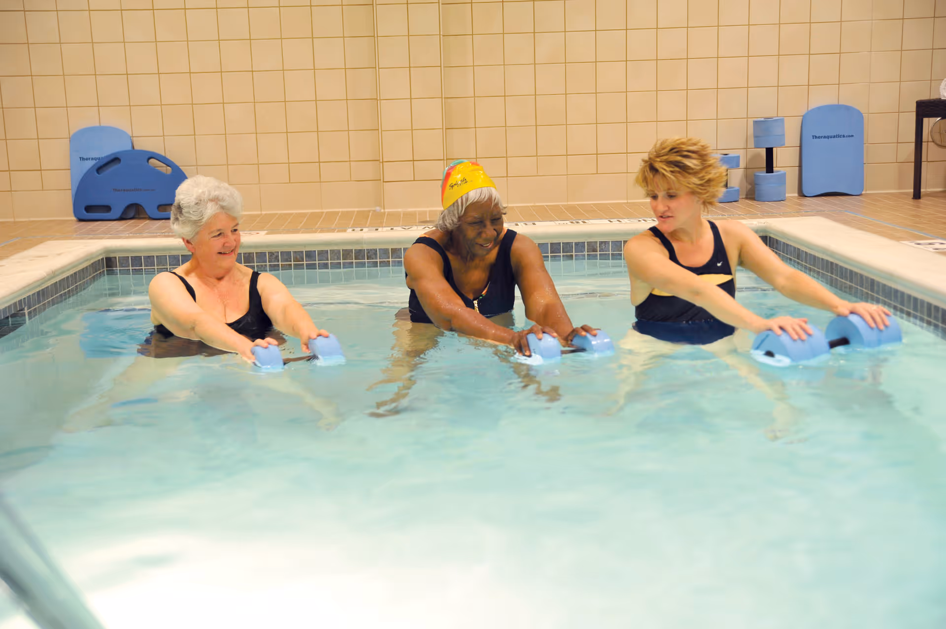 Three women in swimsuits participating in a water exercise class in an indoor pool, each holding blue foam dumbbells and smiling.