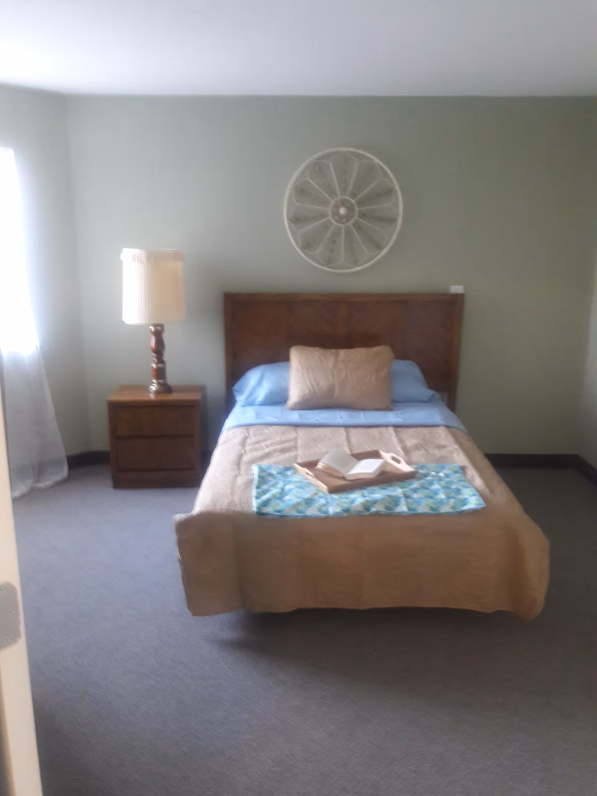 A simple bedroom with a single bed covered with a beige quilt and a blue sheet. There is a beige pillow and a wooden tray with an open book on the bed. A wooden nightstand with a lamp is positioned to the left of the bed. The walls are painted light green, and a decorative circular wall piece hangs above the headboard. Light enters the room through a window with sheer white curtains on the left side.