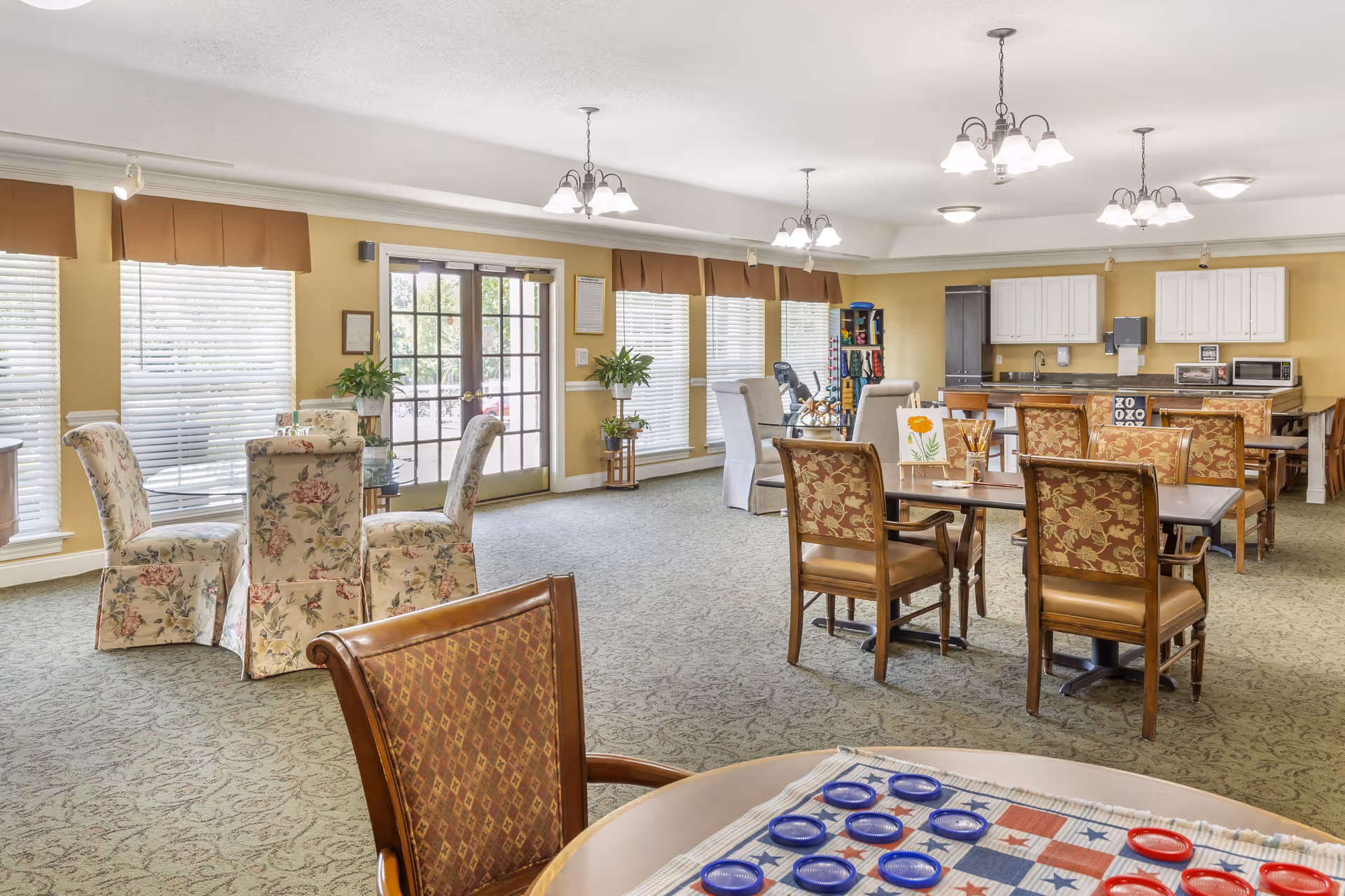 A spacious, well-lit common area in a senior living facility with multiple tables and chairs arranged for social activities. The room features large windows with blinds, floral-patterned chairs, potted plants, and a kitchenette area with white cabinets, a microwave, and a coffee machine. A checkers game is set up on a table in the foreground.