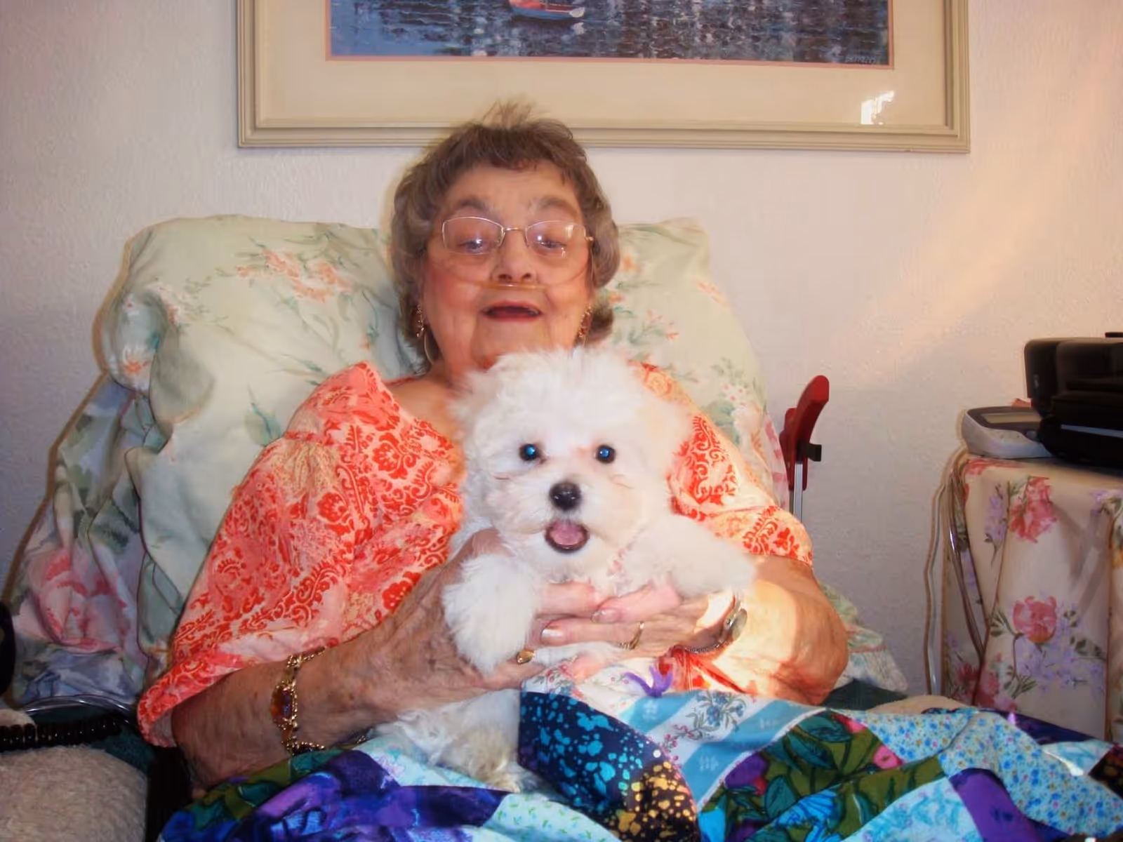An elderly woman sitting in a floral armchair, smiling and holding a small white fluffy dog on her lap. The woman is wearing glasses and a red patterned top, and there is a colorful quilt covering her legs. In the background, there is a framed picture on the wall and a table with a floral tablecloth and some electronic devices.