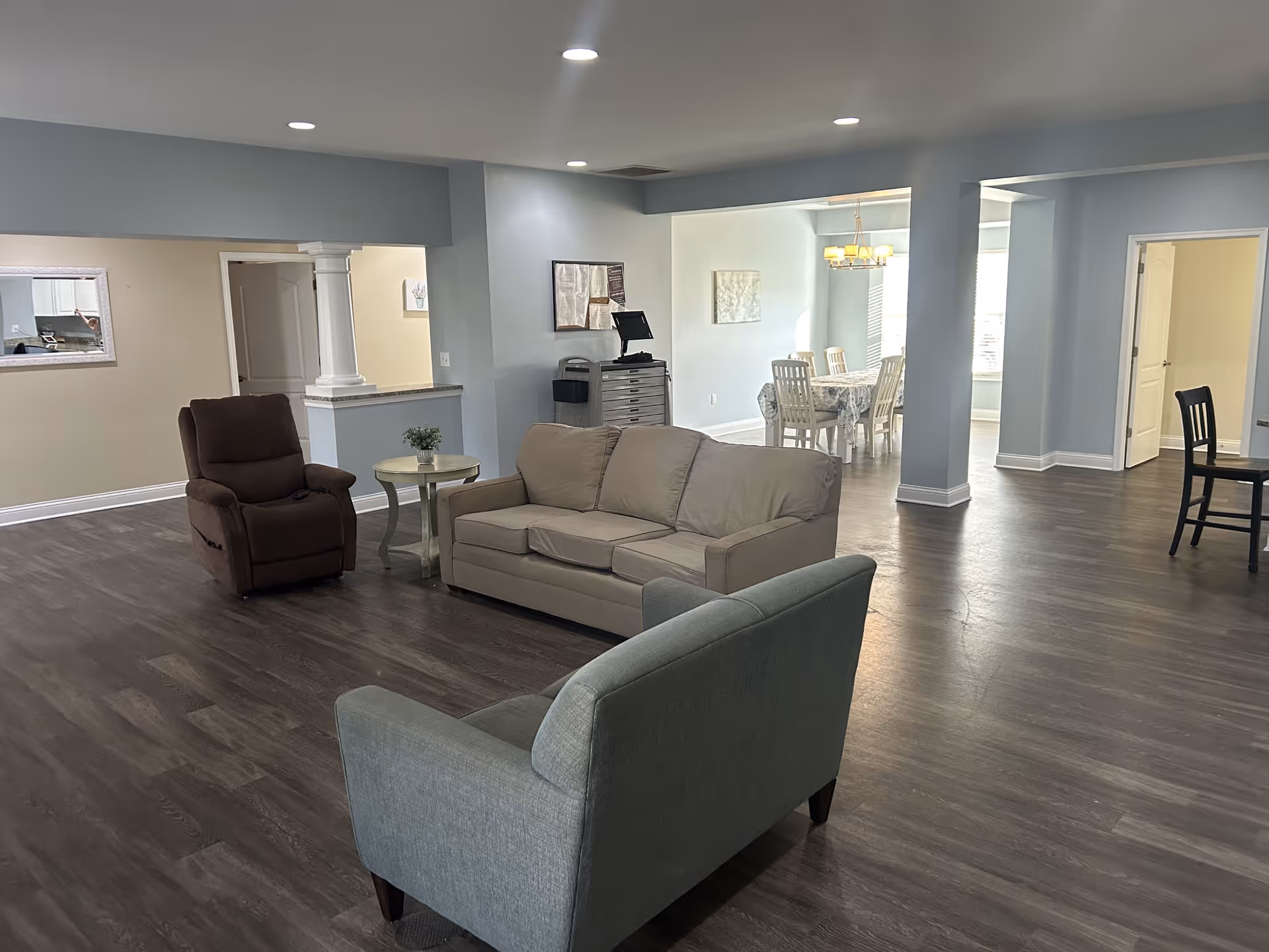 A spacious senior living common area with light blue walls and dark wood flooring. The room features a beige sofa, a brown recliner, a light blue armchair, and a small round side table with a plant. In the background, there is a dining area with a white dining table and chairs under a chandelier. Several doorways and an open layout are visible.