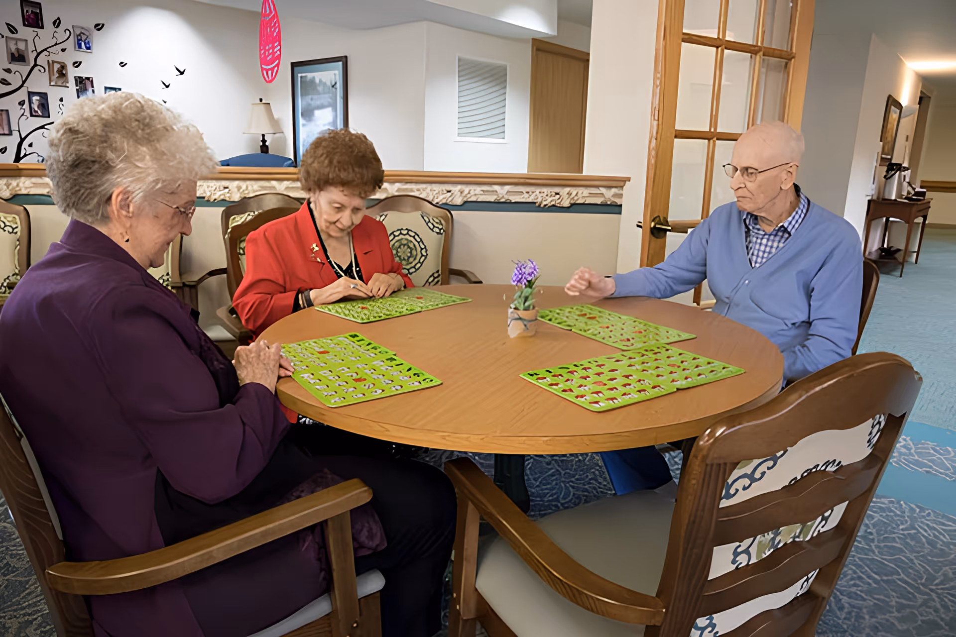 Three elderly individuals sitting around a round wooden table playing a game with green game boards in a well-lit room with decorative chairs and a small potted plant in the center of the table.