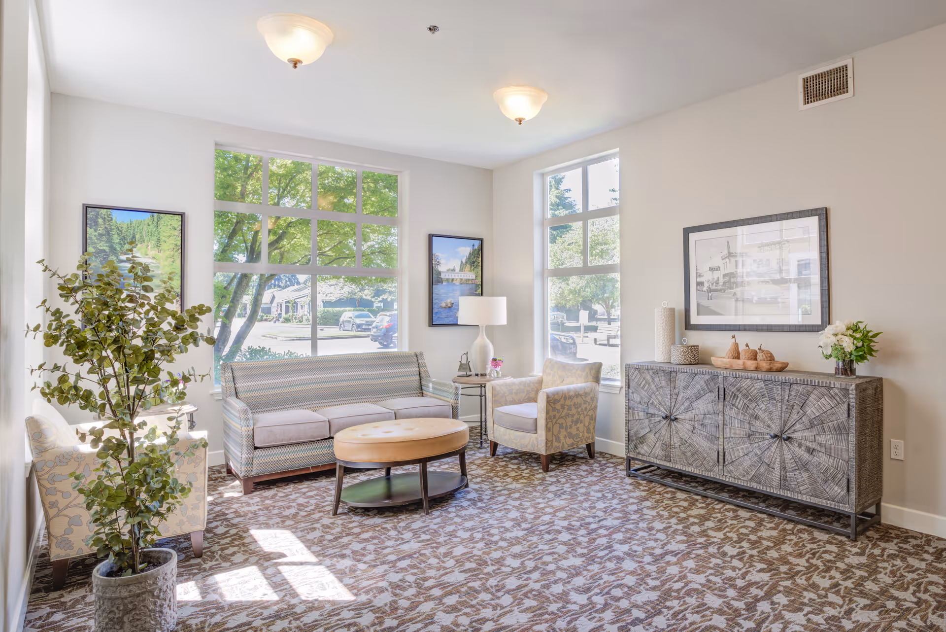 A bright and cozy living room area with large windows letting in natural light. The room features a patterned sofa, two armchairs, a round wooden coffee table, a side table with a lamp, a decorative cabinet with vases and flowers, and framed pictures on the walls. A potted plant is placed near one of the armchairs.