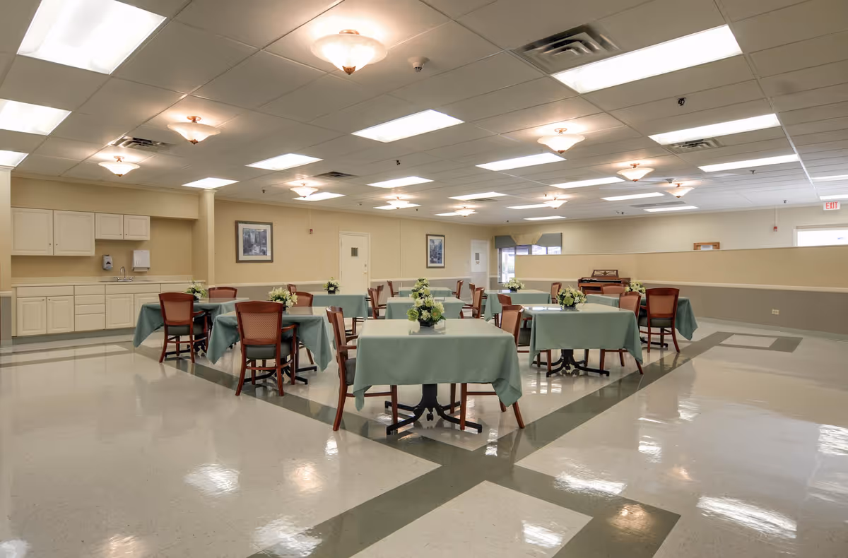 A spacious dining room with multiple square tables covered in green tablecloths, each table surrounded by wooden chairs with red cushions. The tables have floral centerpieces. The room has a tiled floor with a green border pattern, beige walls, and a ceiling with both recessed fluorescent lights and decorative ceiling lights. There is a small kitchenette area with cabinets, a sink, and a soap dispenser on the left side of the room. Two framed pictures hang on the wall, and a piano is visible in the back corner near a window with a valance.