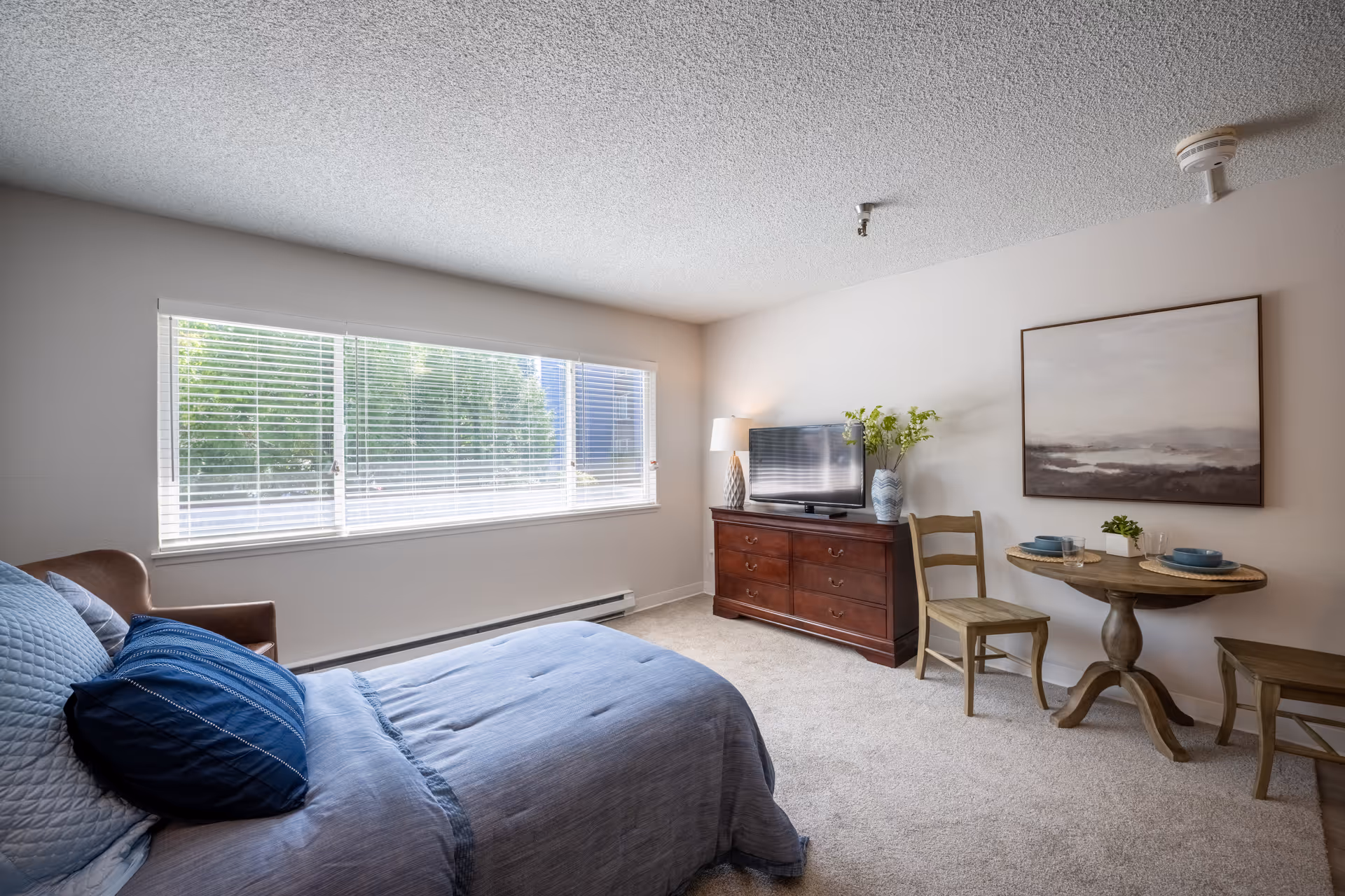 A bright bedroom with a large window covered by white blinds letting in natural light. The room features a bed with blue bedding and pillows, a brown armchair, a wooden dresser with a flat-screen TV, a lamp, and a vase with flowers. There is also a small round wooden table with two chairs, set with plates and glasses, and a large framed landscape painting on the wall.