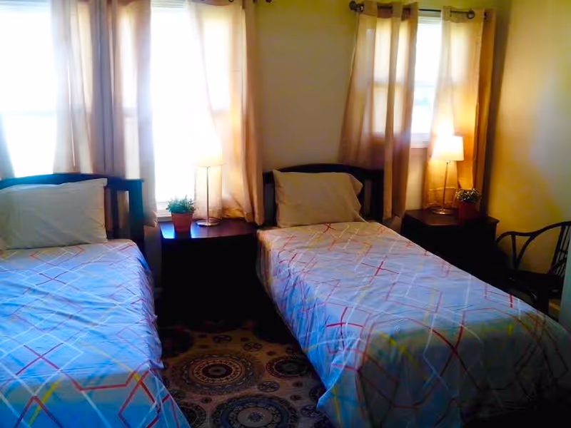 A bedroom with two single beds separated by a nightstand. Each bed has a pillow and a patterned bedspread. There are two windows with beige curtains behind the beds, and two table lamps on the nightstands. A small potted plant is also on each nightstand. A patterned rug covers the floor, and a chair is visible on the right side of the room.