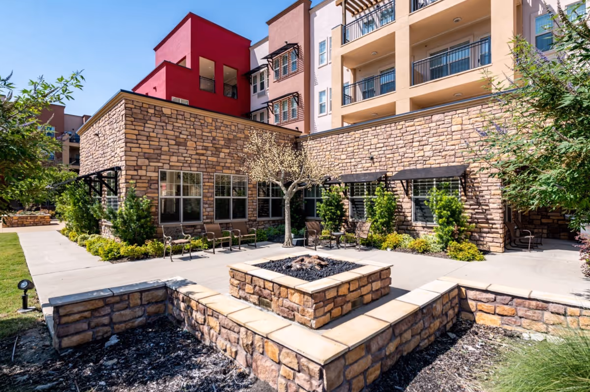 Outdoor courtyard area at Alexis Estates Gracious Retirement Living featuring stone walls, a central fire pit, several chairs arranged around the fire pit, small trees, and shrubs with a multi-story building in the background under a clear blue sky.