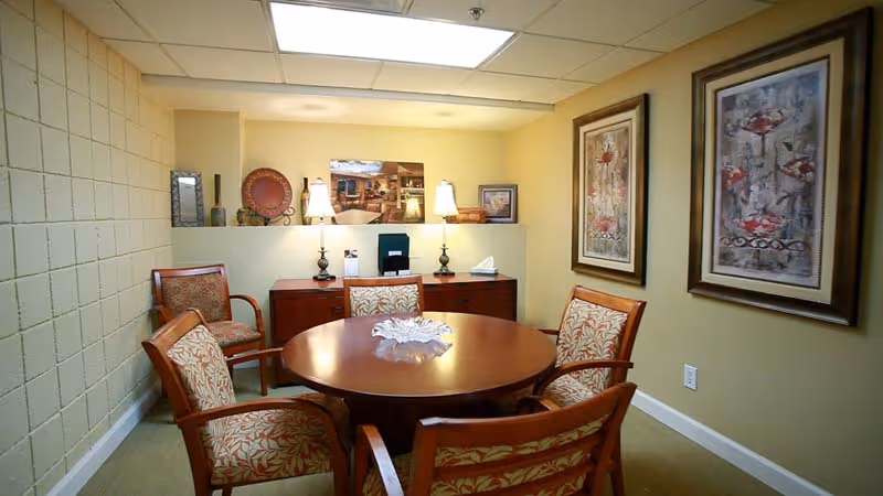 A small meeting or dining room with a round wooden table surrounded by five upholstered chairs with floral patterns. The walls are painted light yellow and decorated with two framed floral paintings. A wooden sideboard against the back wall holds two table lamps, decorative items, and a tissue box. The ceiling has recessed lighting panels.