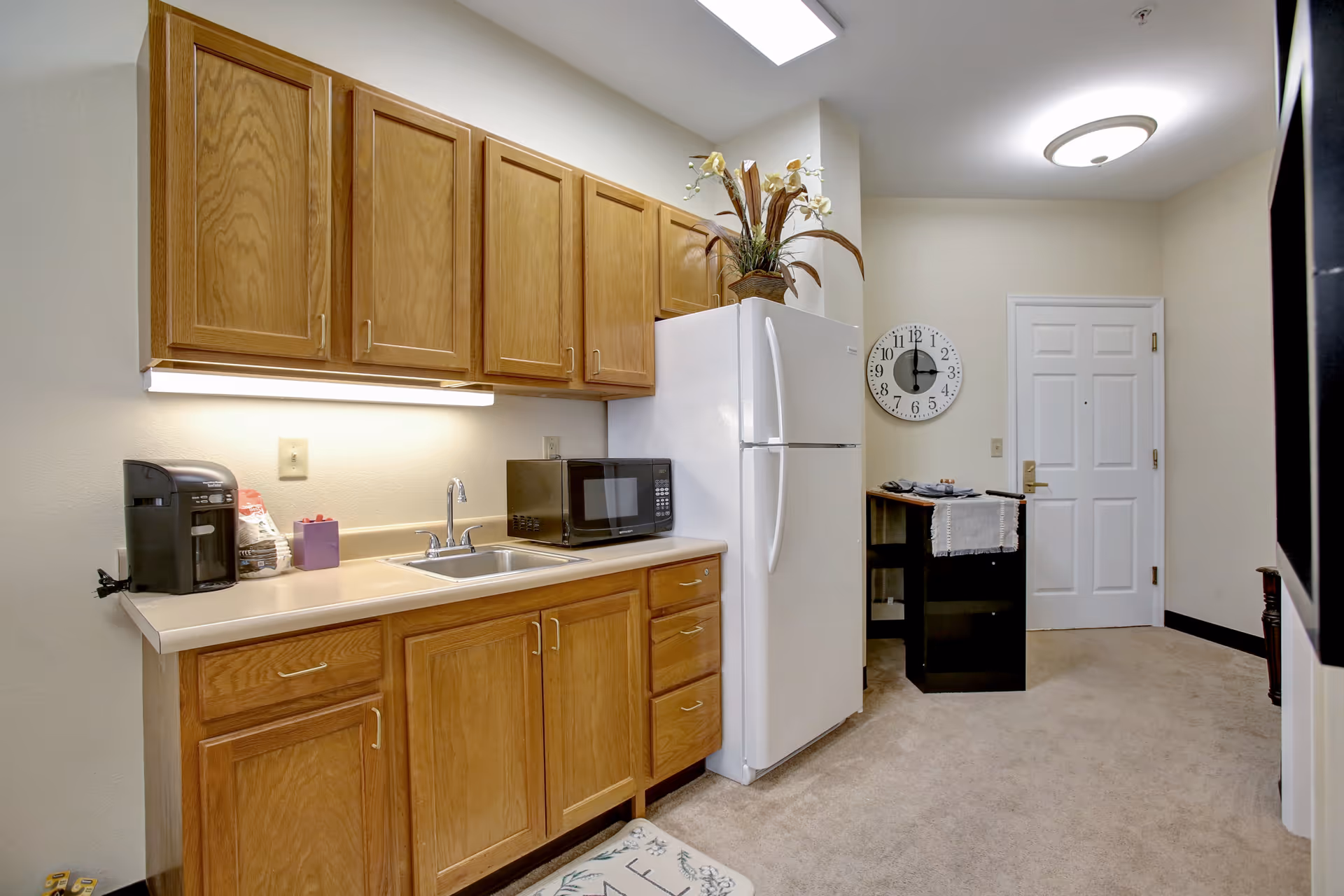 A small kitchen area with wooden cabinets, a white refrigerator, a microwave, a coffee maker, and a sink. There is a decorative plant on top of the refrigerator and a wall clock showing 12:00 above a small black table with folded clothes near a white door. The floor is carpeted.