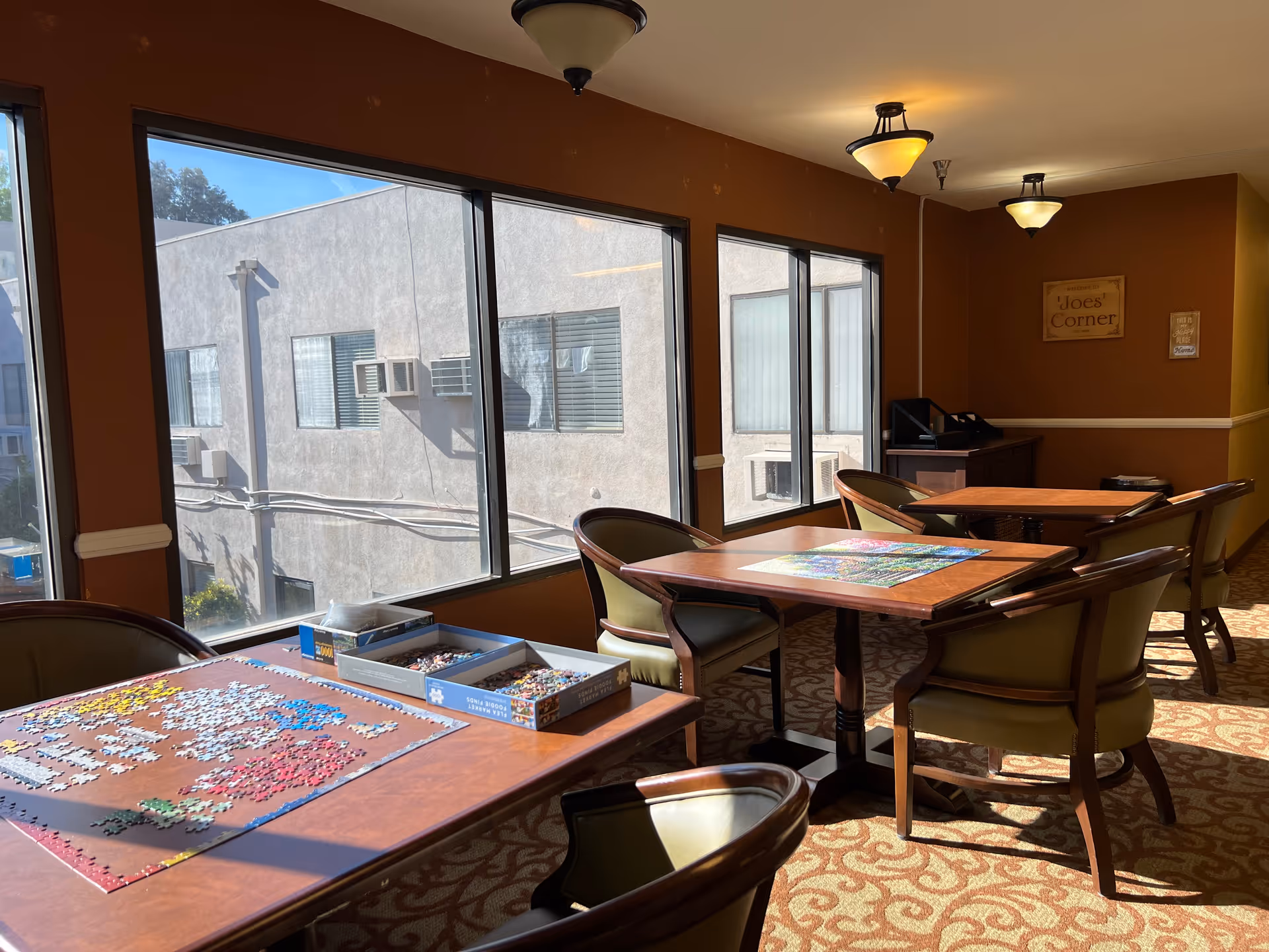 A sunlit room with large windows showing the exterior of a building. Inside, there are several wooden tables with green cushioned chairs around them. On one table, there is a partially completed jigsaw puzzle with puzzle pieces and boxes. The walls are painted brown, and there are ceiling lights. A sign on the wall reads 'Joe's Corner'.