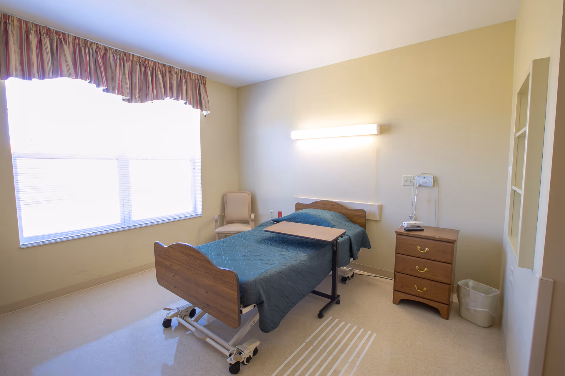 A simple, clean bedroom in a senior living facility with a single hospital-style bed covered with a teal quilt. There is a wooden bedside table with three drawers, a beige armchair near a large window with blinds and a striped valance, a wall-mounted light above the bed, and a small overbed table positioned on the bed.