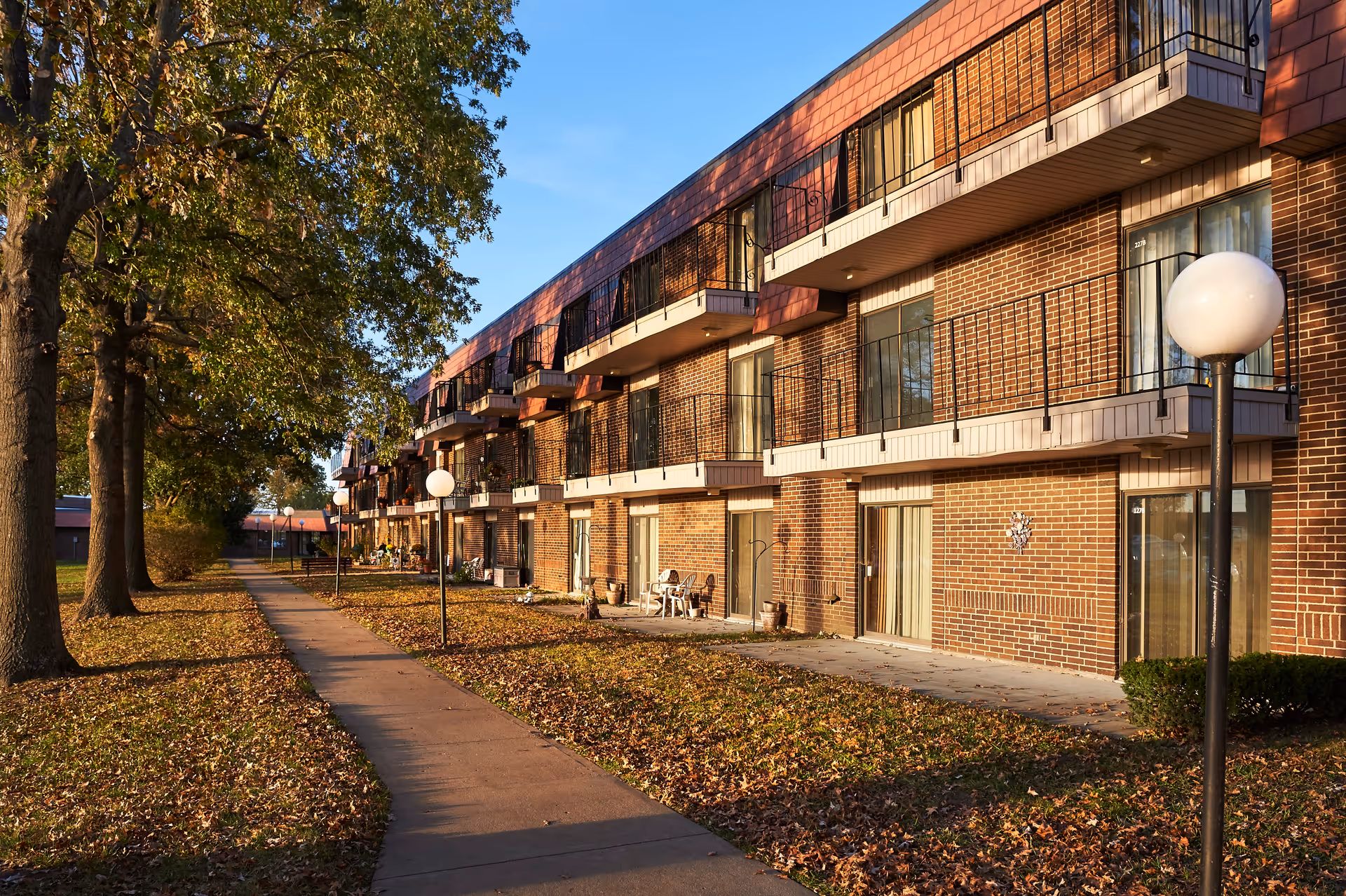 Exterior view of a multi-story brick building with balconies and sliding glass doors, adjacent to a sidewalk lined with trees and lamp posts, with fallen leaves on the ground during autumn.