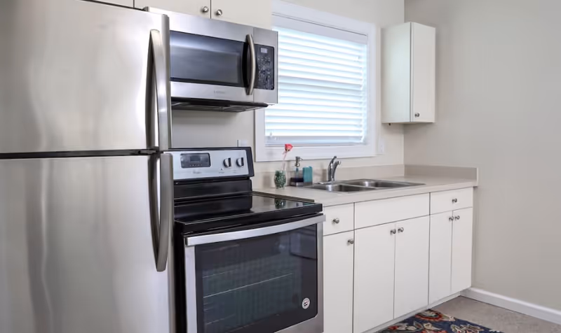 A clean kitchen area featuring stainless steel appliances including a refrigerator, microwave, and electric stove with oven. White cabinets and drawers are below the countertop and a double sink is positioned under a window with white blinds. A small decorative flower and soap dispenser are on the counter.