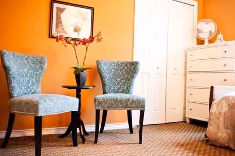 Two blue upholstered chairs flank a small table with a vase of flowers on orange wall