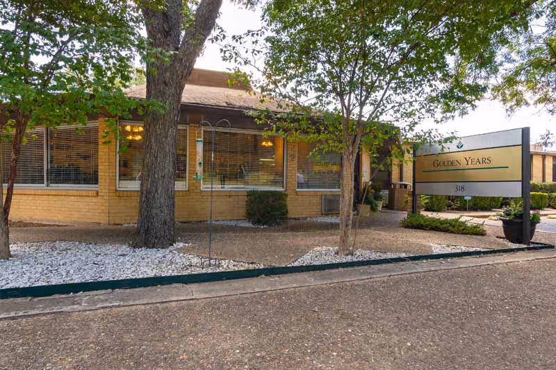 Exterior view of Golden Years Nursing and Rehab facility showing a single-story brick building with large windows, trees, and a sign with the facility name and address 318. The area around the building has landscaping with white rocks and bushes.