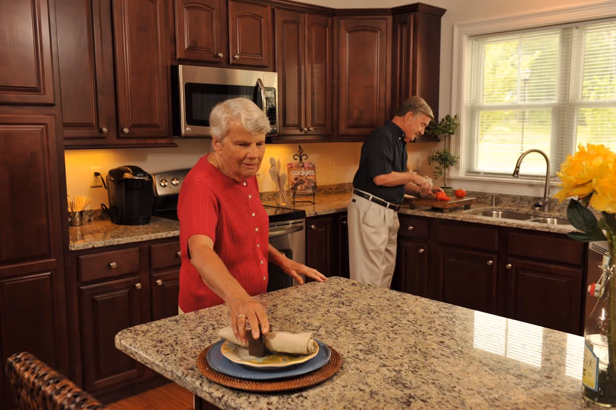 An elderly woman in a red shirt is setting a napkin on a plate on a granite kitchen island, while an elderly man in a dark shirt and light pants is cutting tomatoes on a wooden cutting board near a window in a kitchen with dark wooden cabinets and stainless steel appliances.