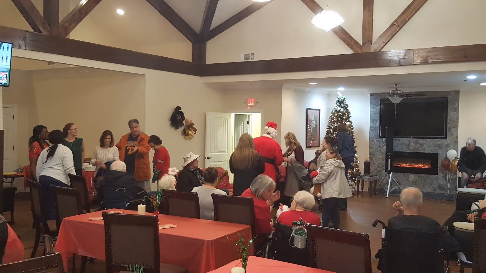 A group of elderly residents and visitors gathered in a senior care home common area decorated for Christmas, featuring a Christmas tree, a fireplace, and a person dressed as Santa Claus interacting with the attendees.