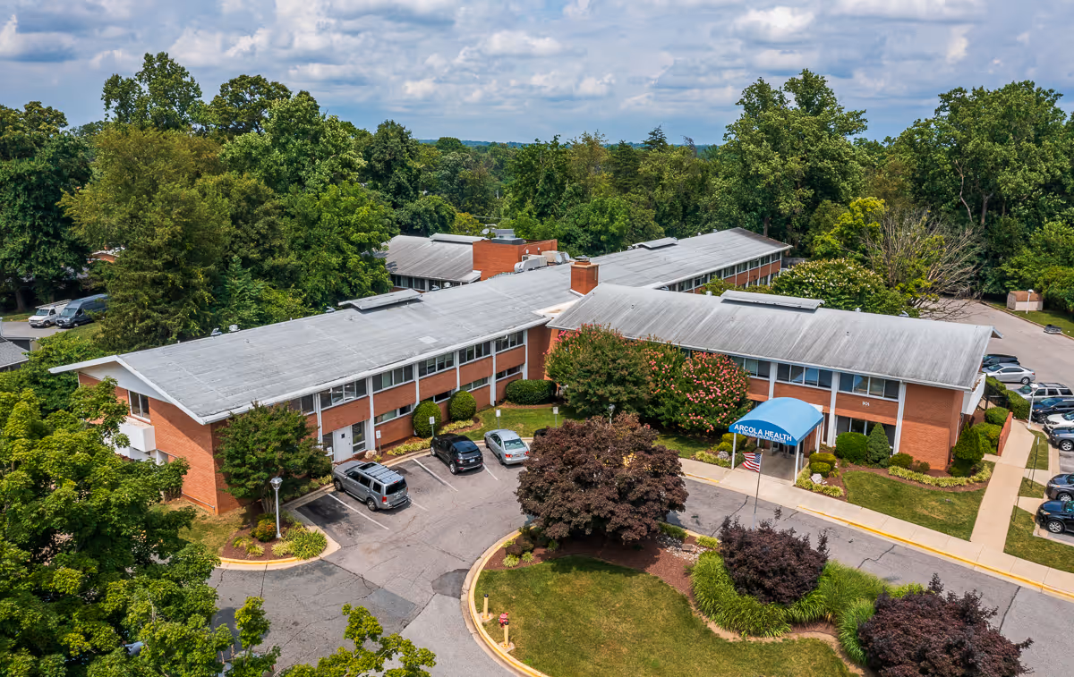 Aerial view of a two-story brick healthcare facility surrounded by trees and greenery. The building has a gray roof and a blue awning at the entrance with the text 'ARCOLA HEALTH'. Several cars are parked in the parking lot around the building.