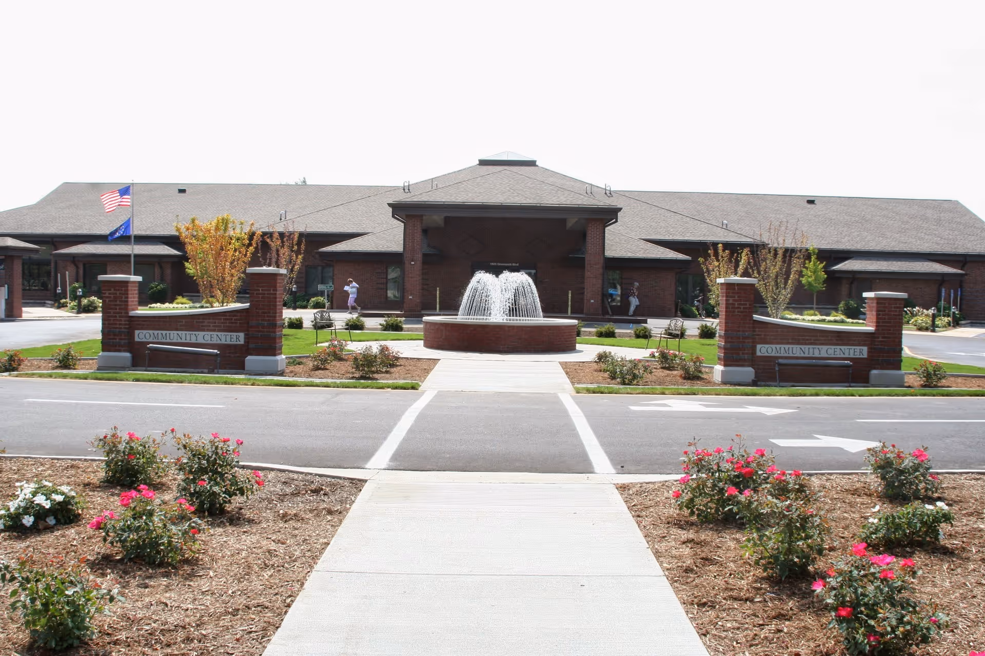 Front exterior of a community center building with a central fountain, brick pillars, flags and landscaped flower beds.
