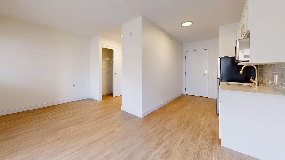 Empty interior space with light wood flooring and white walls. On the right side, there is a small kitchen area with white cabinets, a sink, a microwave, and a refrigerator. In the center, there is a partial wall dividing the space. On the left side, there is an open closet with a sliding door. A white entrance door is visible at the far end of the room.
