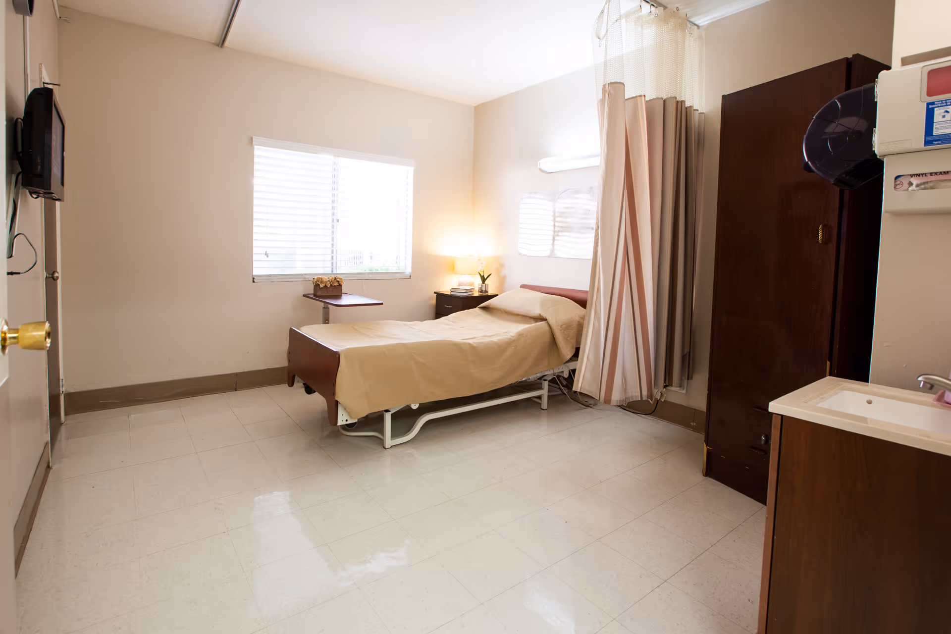 A sparse single-occupancy care room with a hospital-style bed, nightstand and lamp, privacy curtain, wardrobe, sink, and wall-mounted TV.