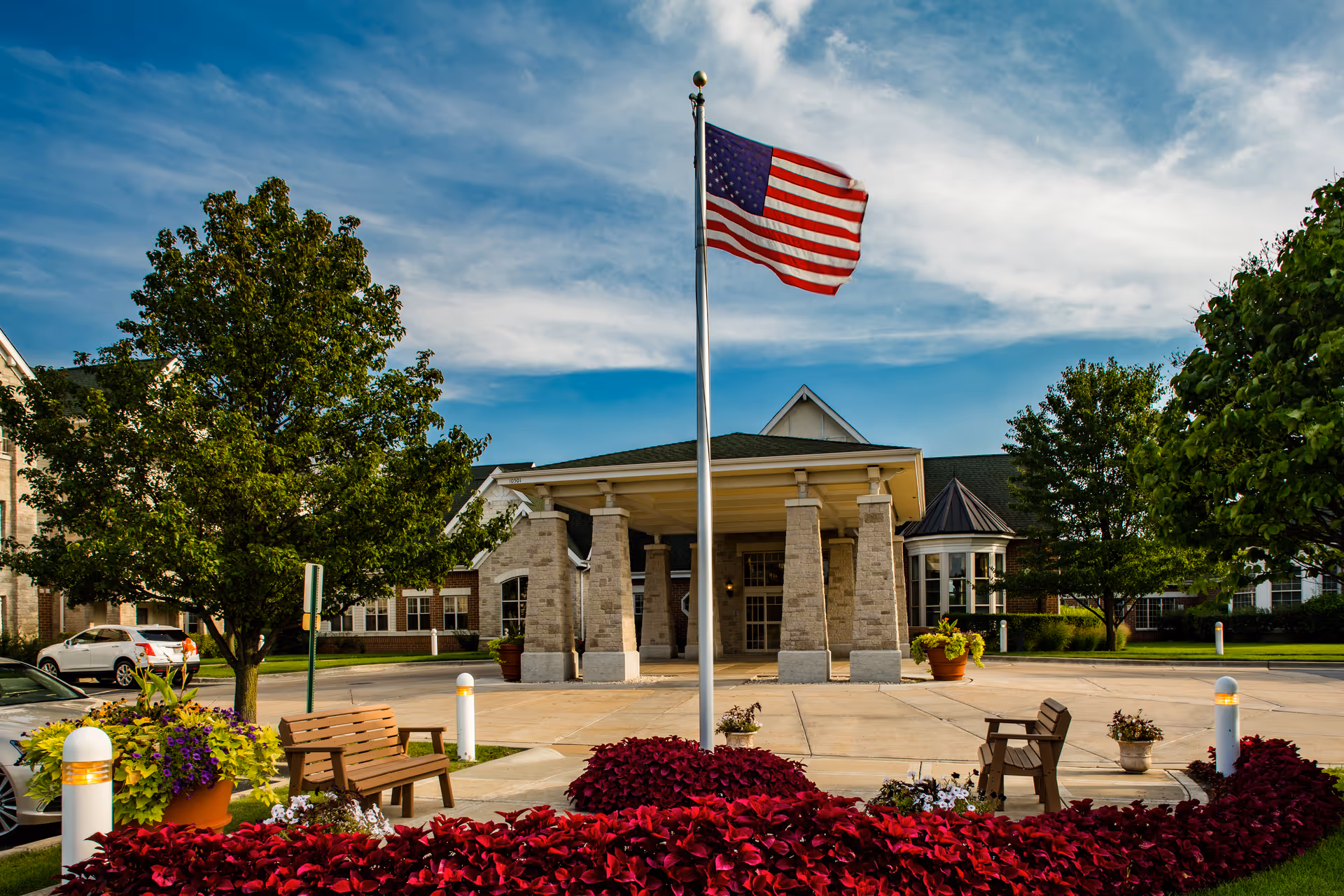 Entrance of a senior living facility with a covered driveway supported by stone pillars, an American flag on a flagpole in the center, surrounded by trees, benches, flower pots, and a well-maintained garden under a partly cloudy sky.