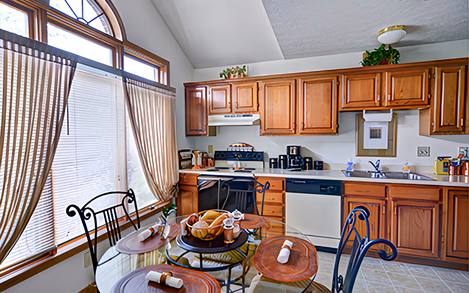 A bright kitchen with wooden cabinets, a glass dining table set with four chairs, and large windows with sheer curtains allowing natural light to fill the room. The kitchen features a stove, dishwasher, sink, and various small appliances on the countertop. A decorative fruit bowl is placed on the dining table.