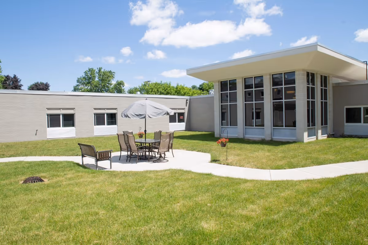 Outdoor courtyard area of a senior living facility with green grass, a concrete walkway, a patio table with six chairs and an umbrella, and a bench. The building has large windows and a flat roof under a blue sky with some clouds.