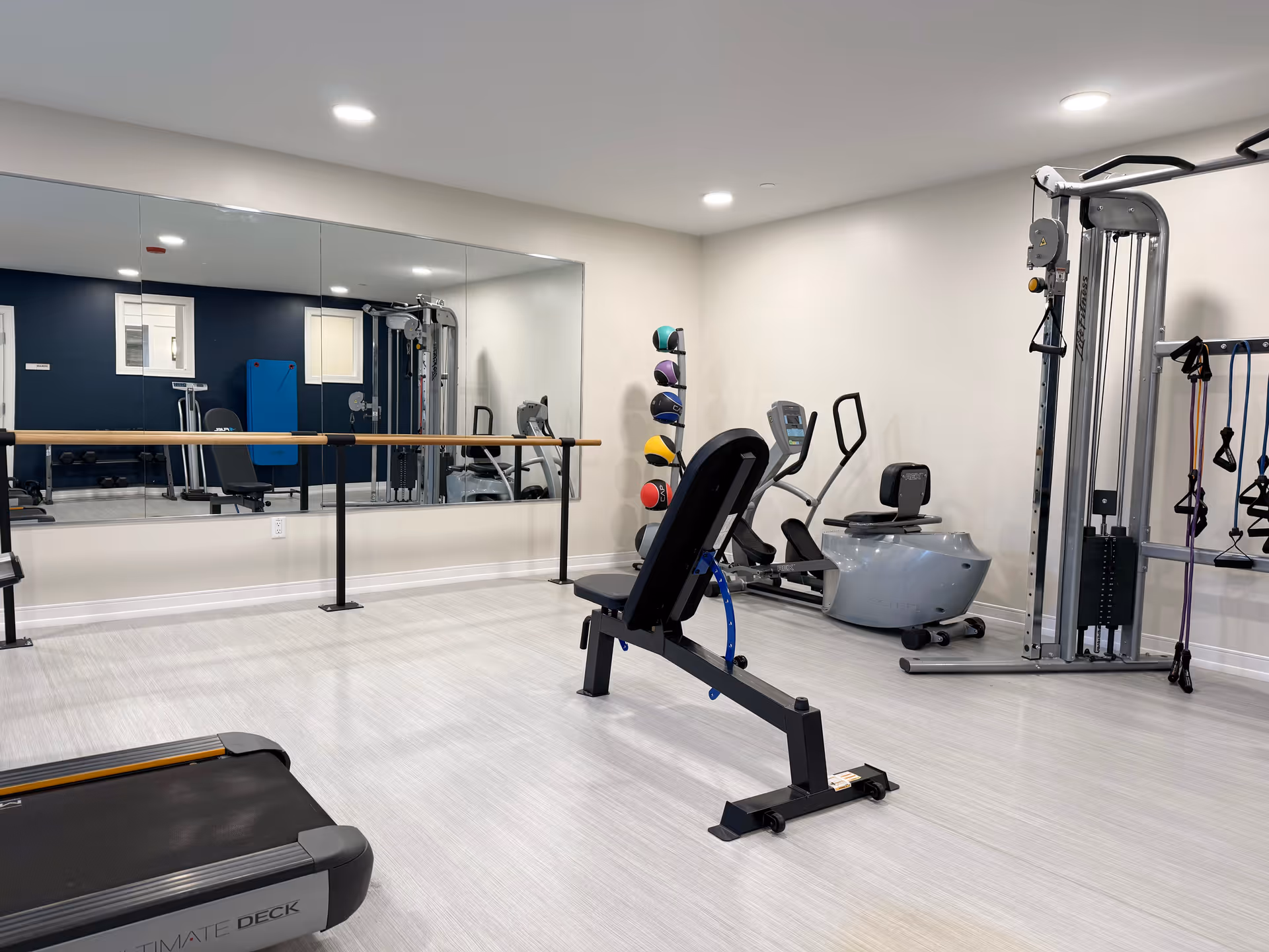 A well-lit fitness room with exercise equipment including a treadmill, adjustable bench, stationary bike, multi-function weight machine, and a rack of colorful medicine balls. A large wall mirror and ballet barre are visible on one side of the room.