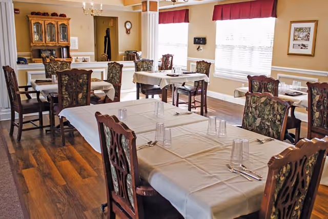 Dining room with multiple tables covered with white tablecloths, set with glasses and silverware. The room has wooden chairs with floral upholstery, hardwood floors, large windows with red valances, and a wooden cabinet against the far wall.