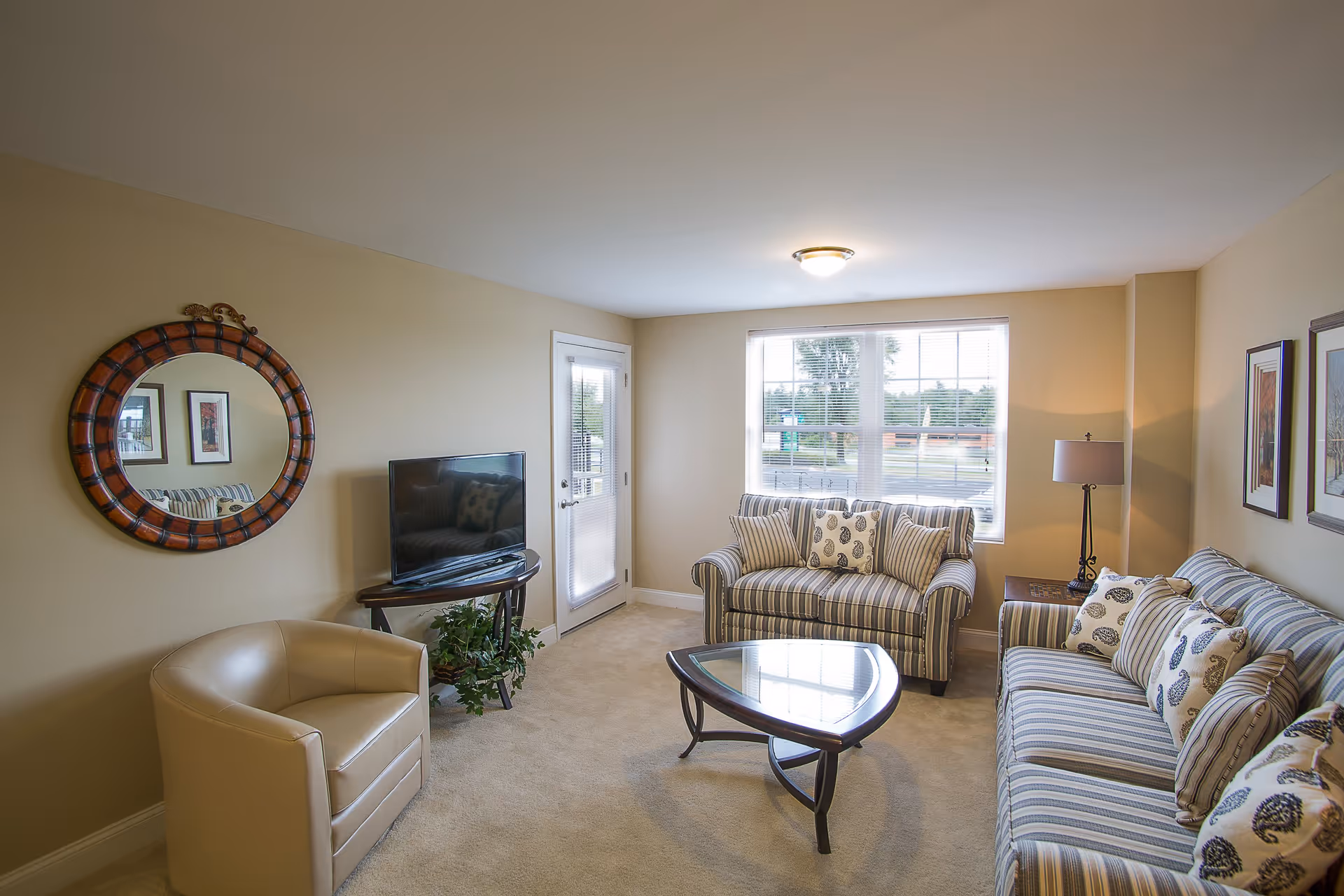 A cozy living room with beige walls and carpet, featuring a striped sofa and loveseat with patterned cushions, a beige armchair, a glass coffee table with a wooden base, a flat-screen TV on a wooden stand, a round decorative mirror on the wall, a floor lamp, and a window letting in natural light.