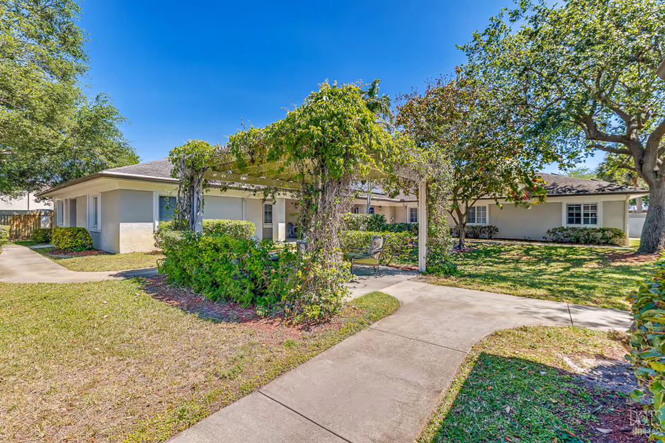 Outdoor garden area at Cottages at Palm Beach featuring a pergola covered with green vines, surrounded by bushes and trees, with a concrete pathway leading through the grassy yard and a beige single-story building in the background under a clear blue sky.