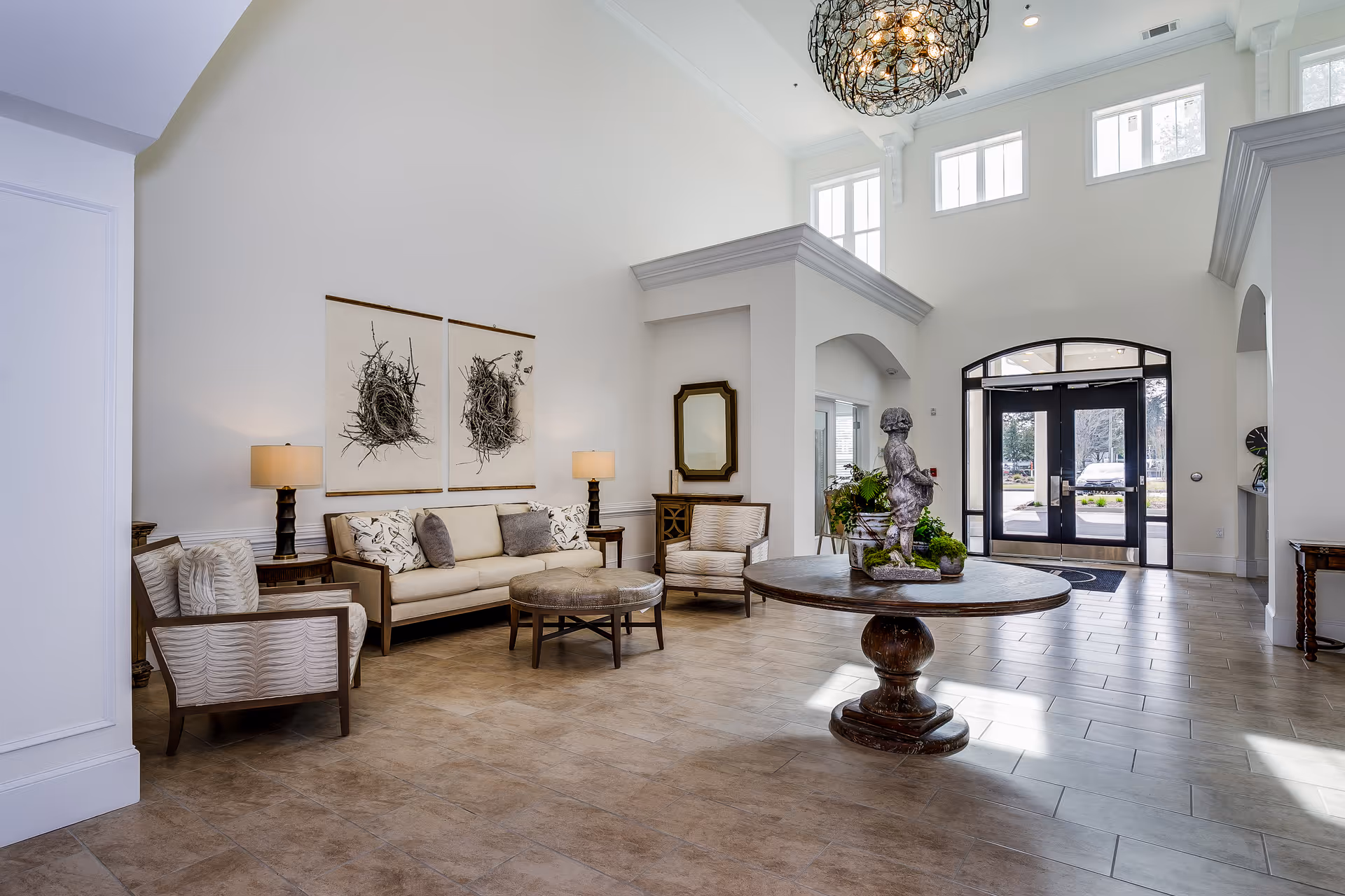 Bright, high-ceilinged senior living lobby with a seating area, round center table topped by a statue and double glass entrance doors.