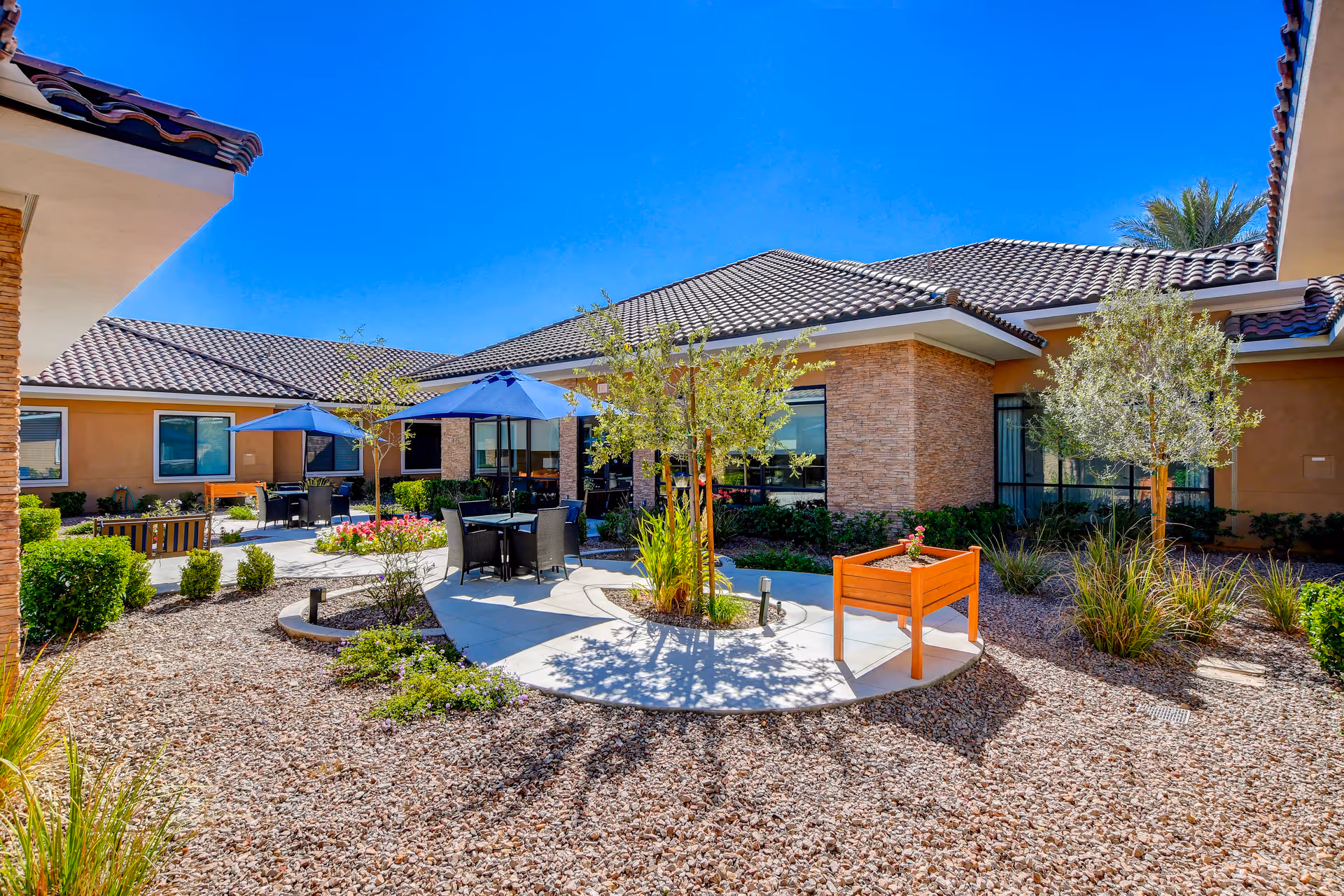 Sunny courtyard with patio tables and umbrellas, raised planters, gravel landscaping, and a surrounding single-story building.