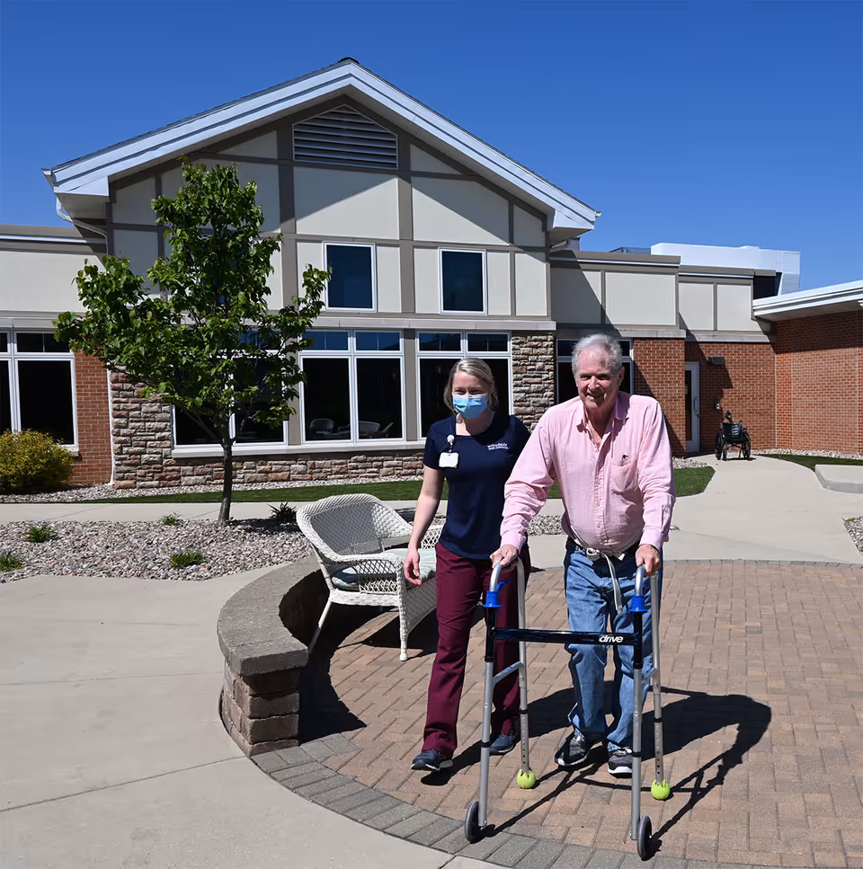 An elderly man using a walker is being assisted by a caregiver wearing a mask outside a building with brick and stone exterior walls under a clear blue sky. There is a small tree, a white wicker chair, and a paved walkway in the courtyard area.