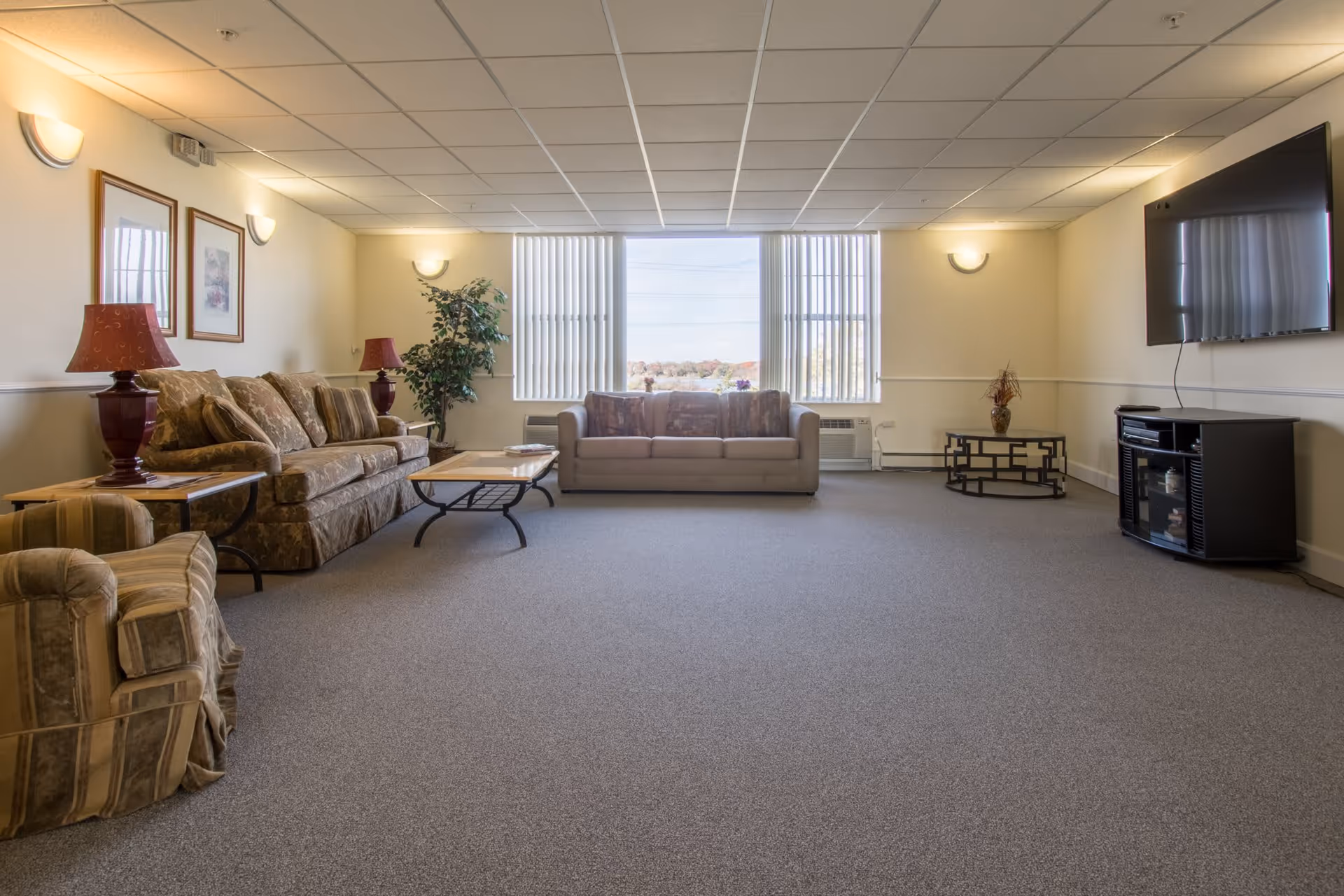 A spacious living room with beige walls and carpeted floor. The room features a patterned sofa, a beige sofa, and a striped armchair. There are two side tables with red lamps, a coffee table, a small round table with a vase, and a flat-screen TV mounted on the wall. Large windows with vertical blinds allow natural light to enter the room.