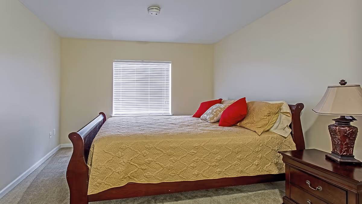 A bedroom with a wooden bed frame and a beige quilted bedspread. The bed has two red pillows and one beige patterned pillow. There is a window with closed blinds on the wall behind the bed. A wooden nightstand with a decorative lamp is positioned to the right of the bed. The walls are painted light beige and the floor is carpeted.