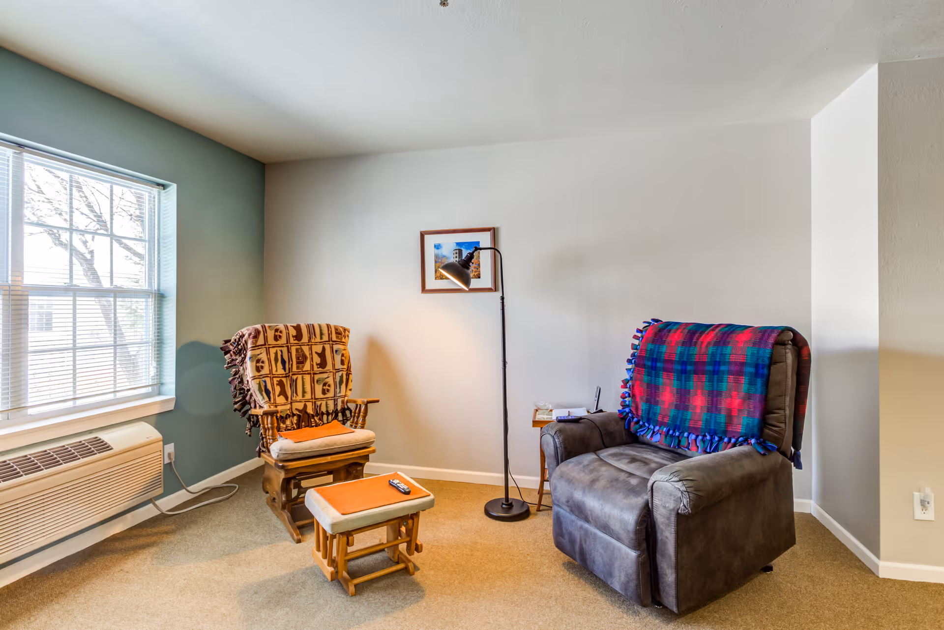 A cozy sitting area with two armchairs, a floor lamp, a window and a small footstool in an assisted living room.