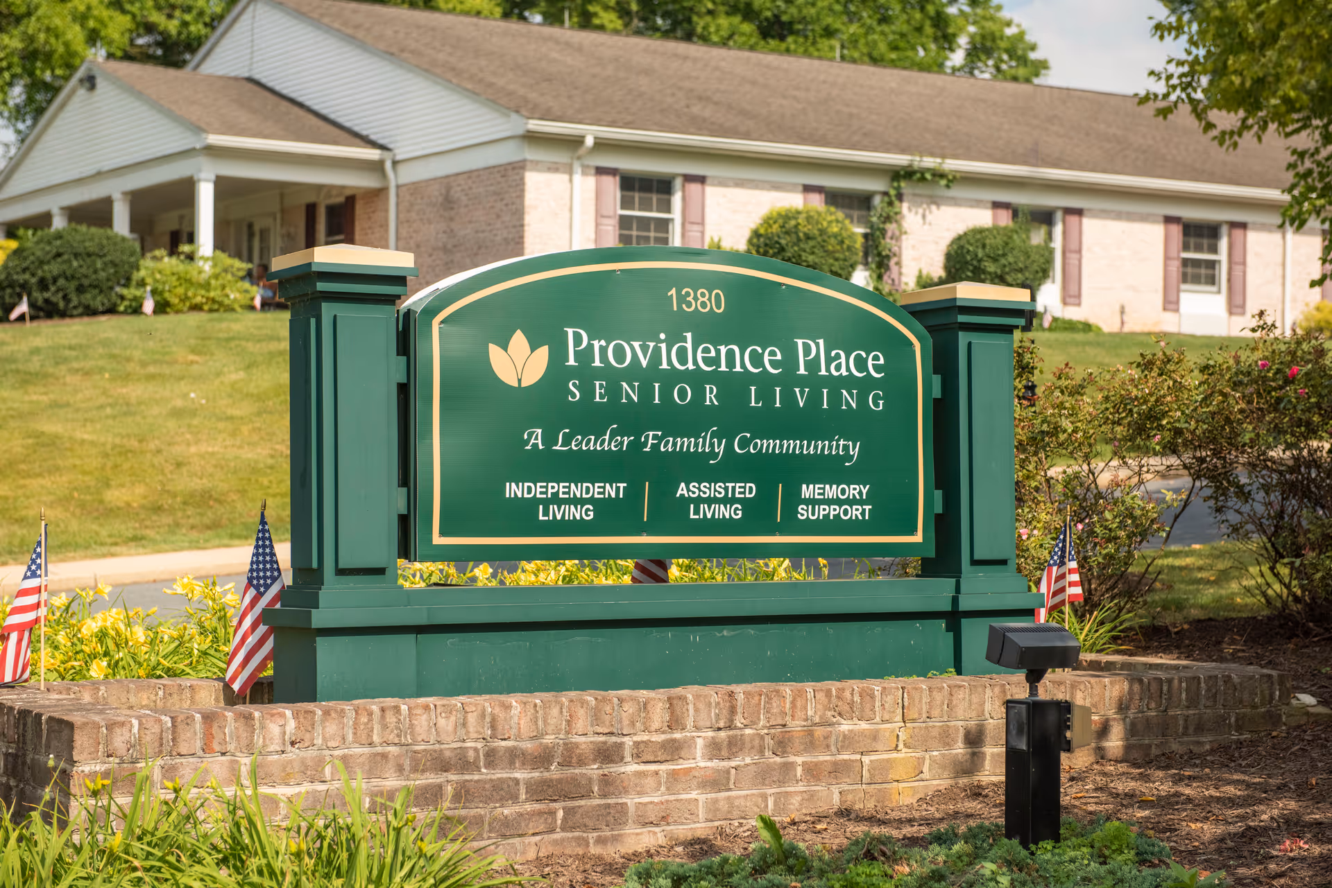 Outdoor view of a green and gold sign for Providence Place Senior Living, located on a brick base with small American flags around it. In the background, there is a single-story building with white siding and a porch, surrounded by greenery and bushes.